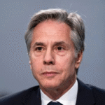Former Secretary of State Antony Blinken gathering his belongings on a beach as a lifeguard stands nearby.